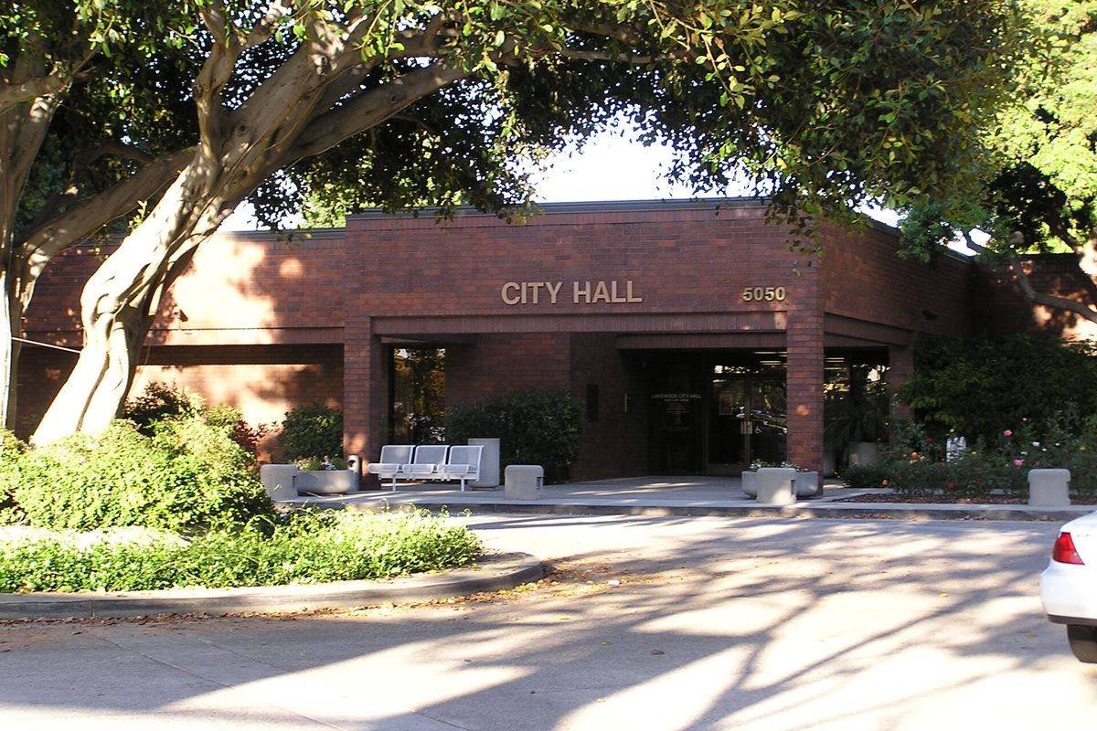 The low brick Lakewood City Hall building with City Hall in raised letters over the entrance, shaded by a large tree, with benches out front