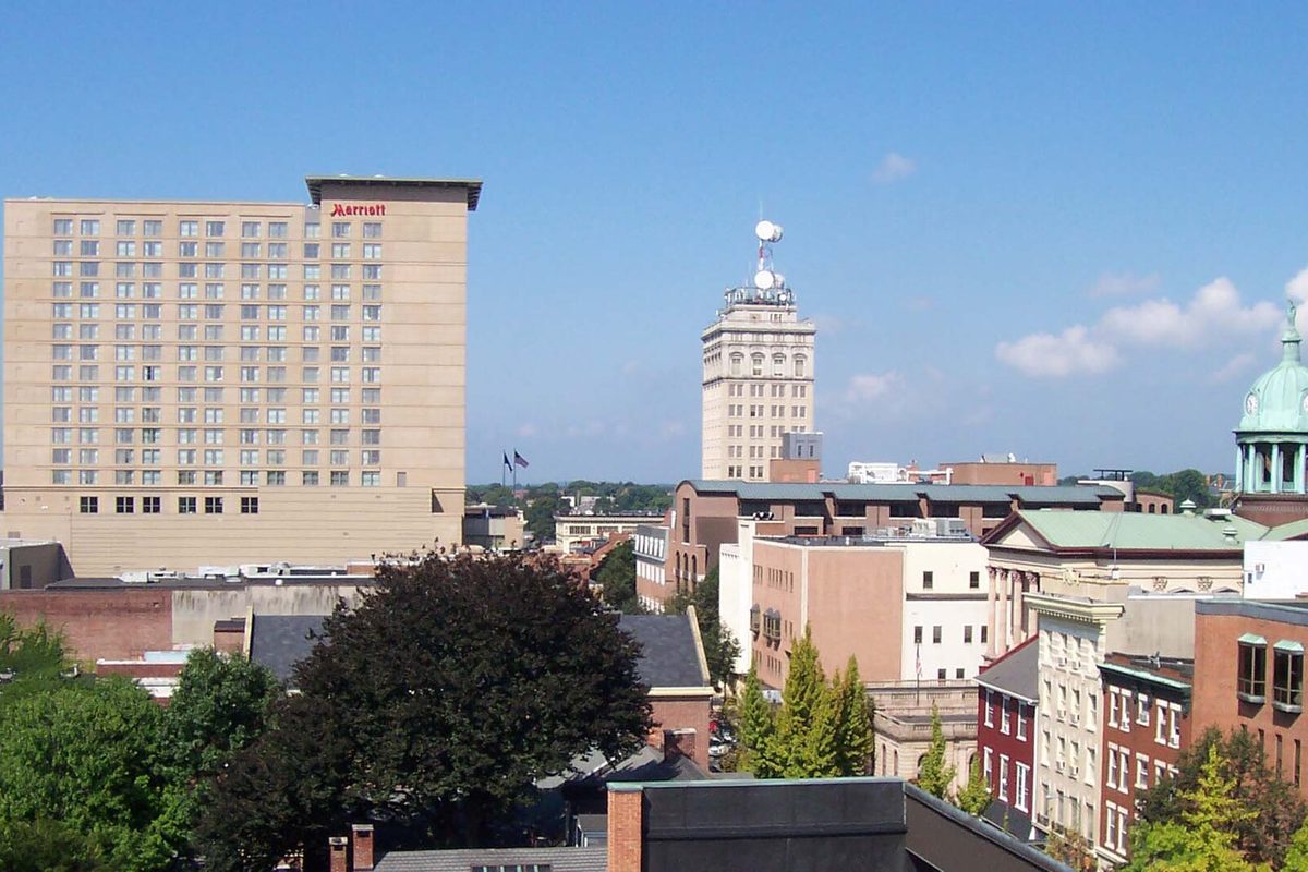 Downtown Lancaster rooftops under a clear blue sky, with a tall tan hotel at the left, an older stepped office tower in the middle and a green dome at the right edge