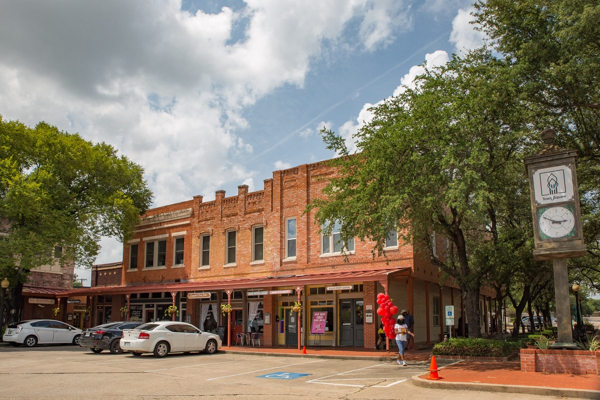 Two-story red-brick storefronts on Lancaster's downtown square with a street clock, shade trees, red balloons and parked white cars under a cloudy sky