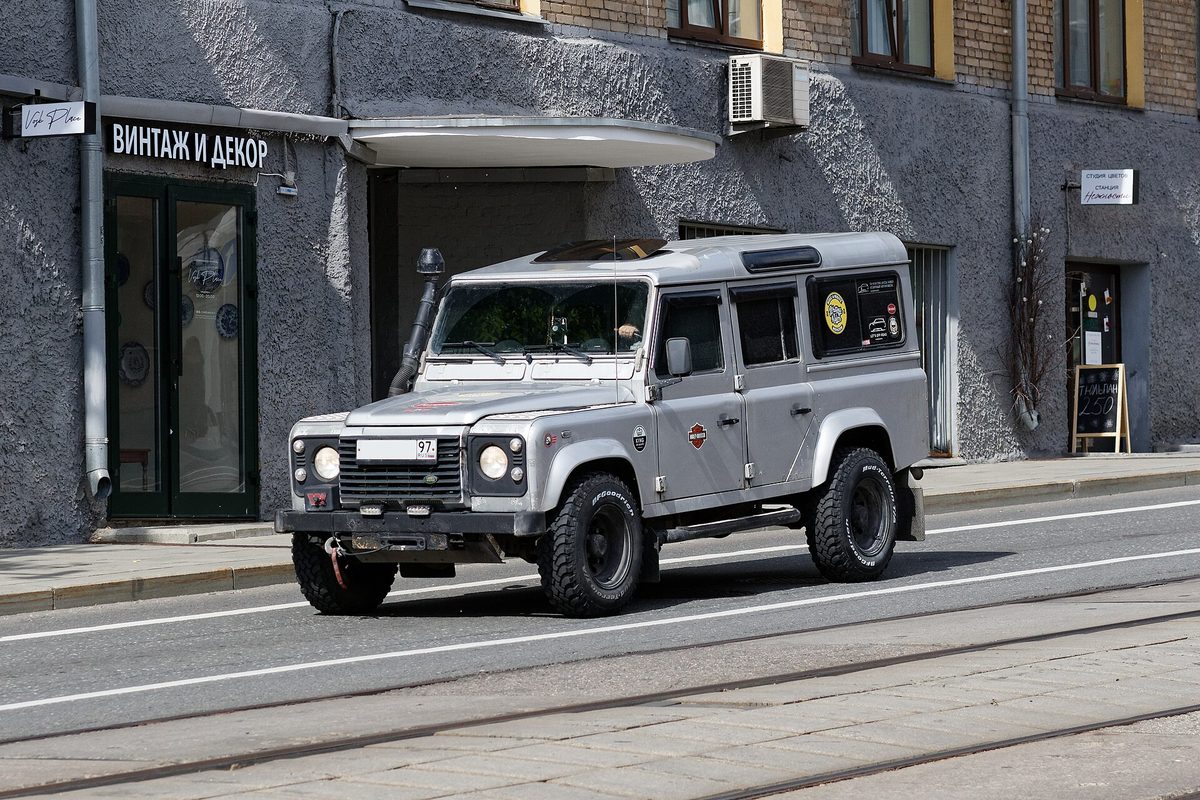 A silver five-door Land Rover Defender with a snorkel and off-road tires, driving past a gray building on a city street with tram tracks