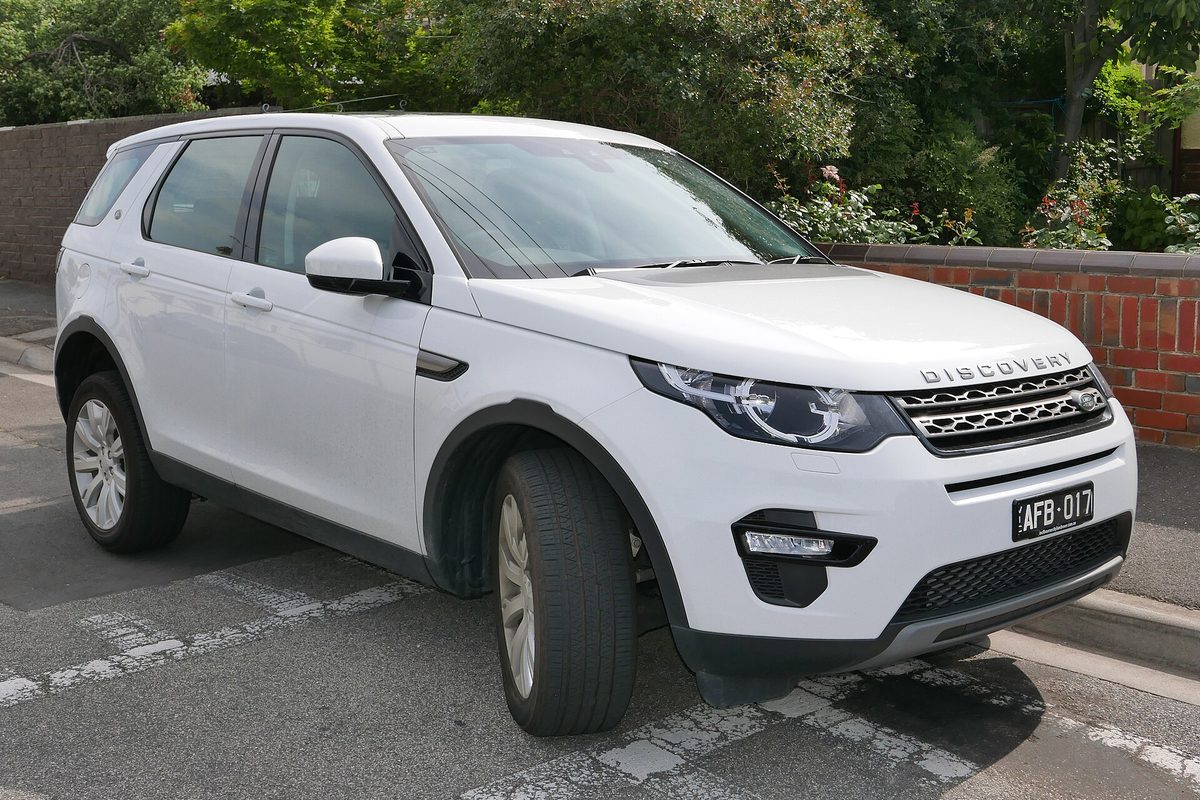 A white 2015 Land Rover Discovery Sport parked at a street curb in front of a brick wall and green hedges