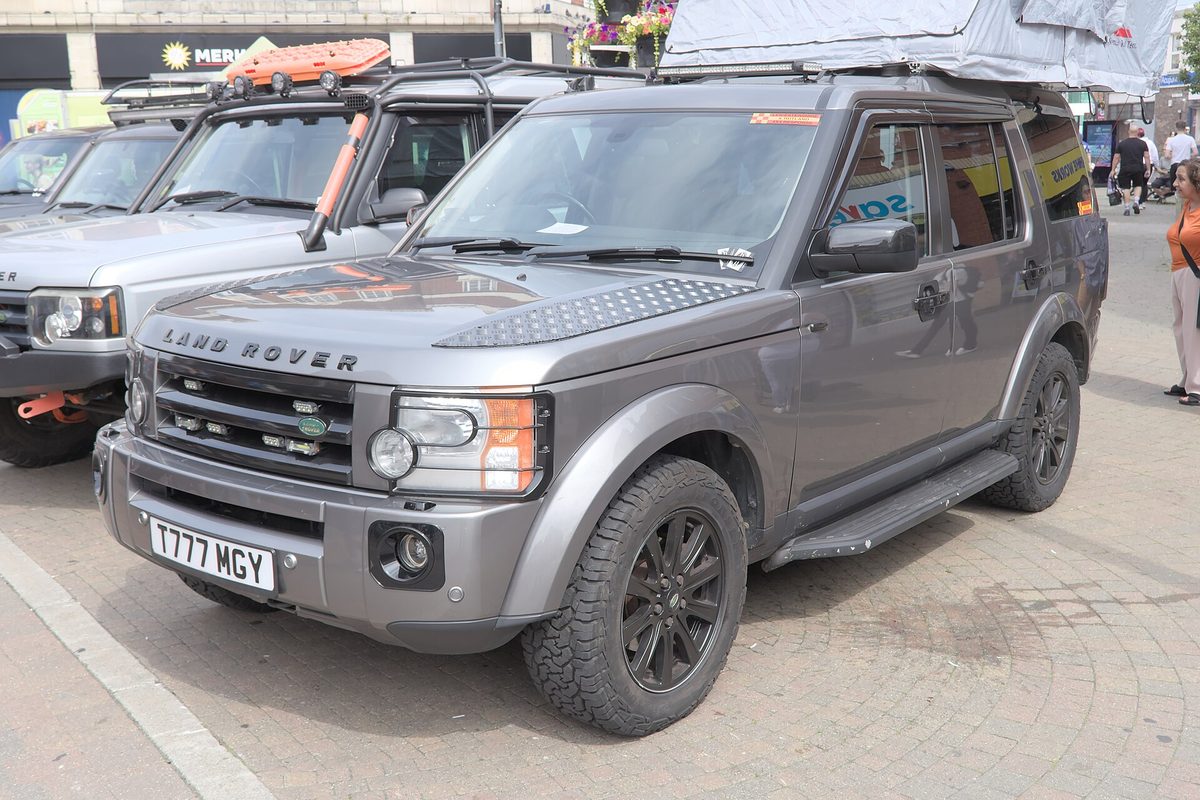 A gray Land Rover Discovery with black wheels, side steps and a loaded roof rack, parked at an outdoor car show in a town square
