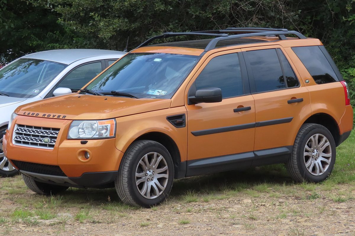 An orange 2008 Land Rover LR2 SE with roof rails, parked on patchy grass next to a silver car, with trees behind it