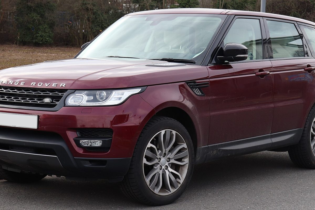 A dark red Range Rover Sport seen from the front left corner, parked on a road beside a bare hedge on a gray day