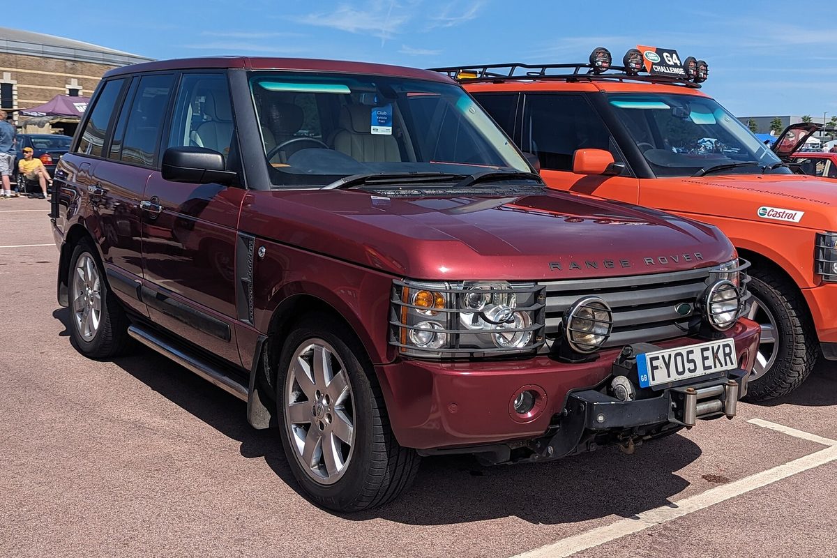 A dark red Range Rover with a grille guard and extra driving lights, parked on a sunny show field next to an orange off-roader
