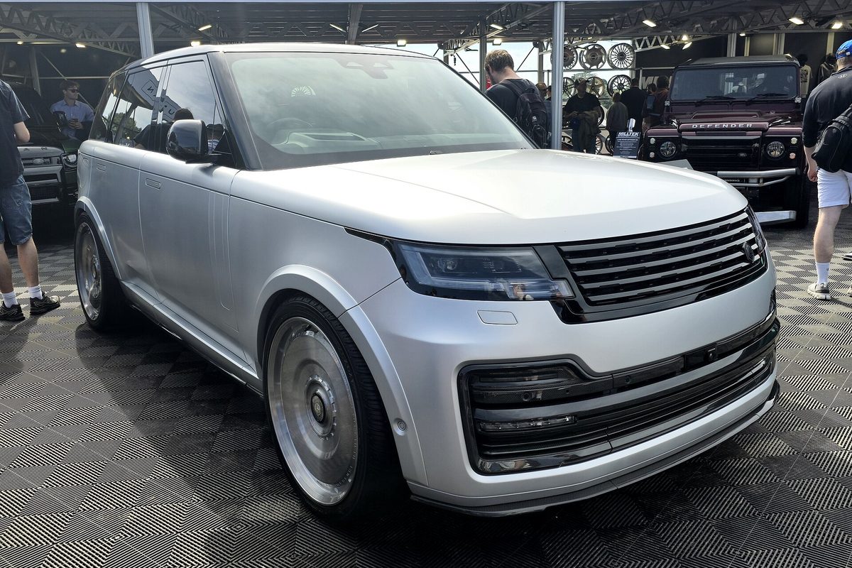 A silver 2024 Range Rover on large custom wheels, parked on a checkered floor at an outdoor car show, with a dark red Defender behind it