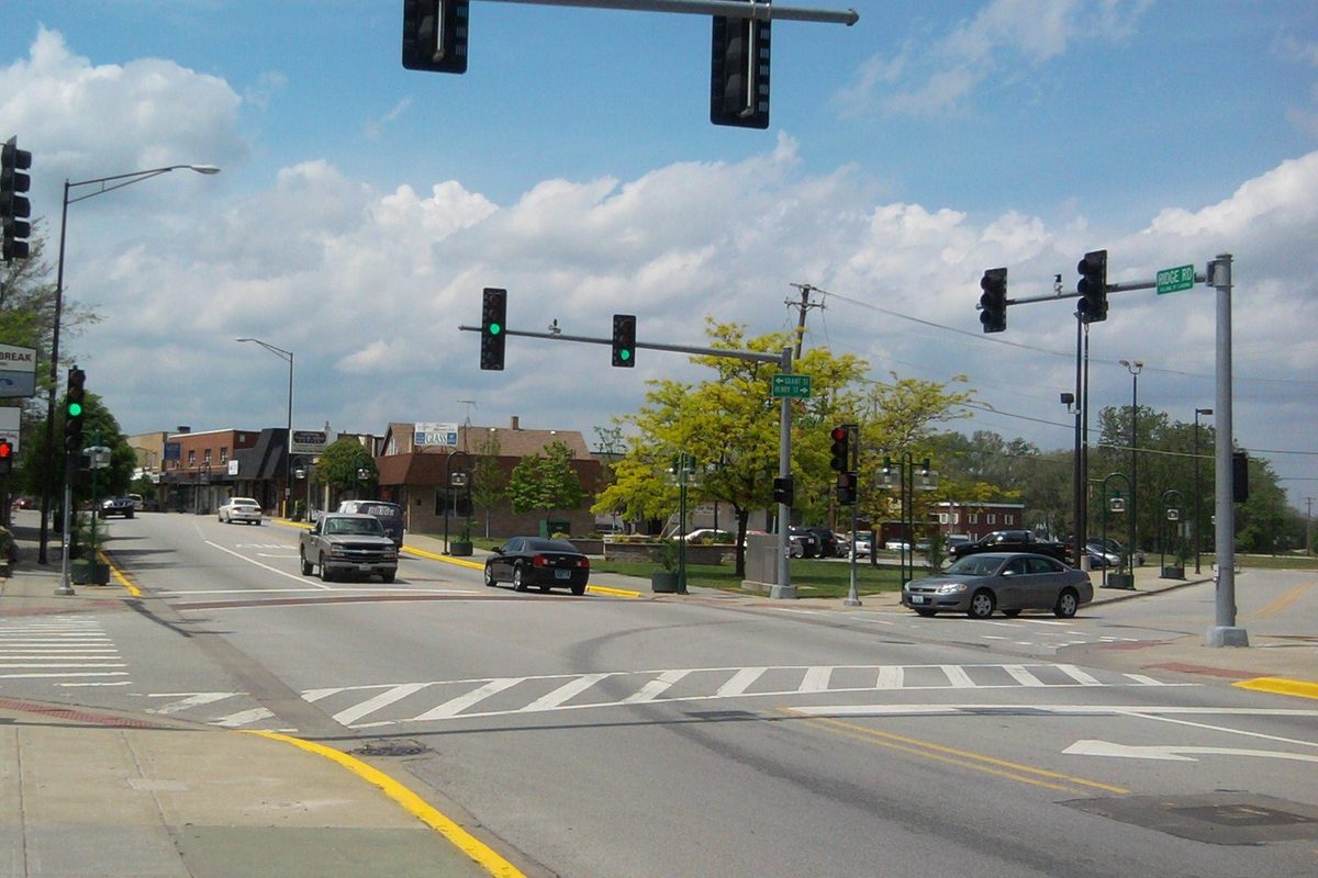 A wide intersection on Ridge Road in downtown Lansing with traffic signals overhead, low brick storefronts and a few cars under a cloudy sky