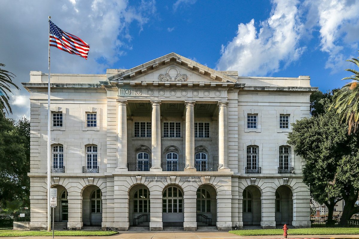 The white stone U.S. Post Office, Court House and Custom House in Laredo, with tall columns, arched ground-floor windows, an American flag and palm trees under a blue sky