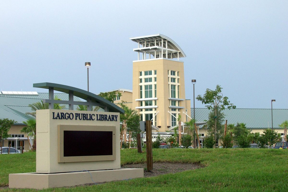 The Largo Public Library sign on a grassy lawn, with the library's tall glass-and-stucco tower and green metal roof behind it