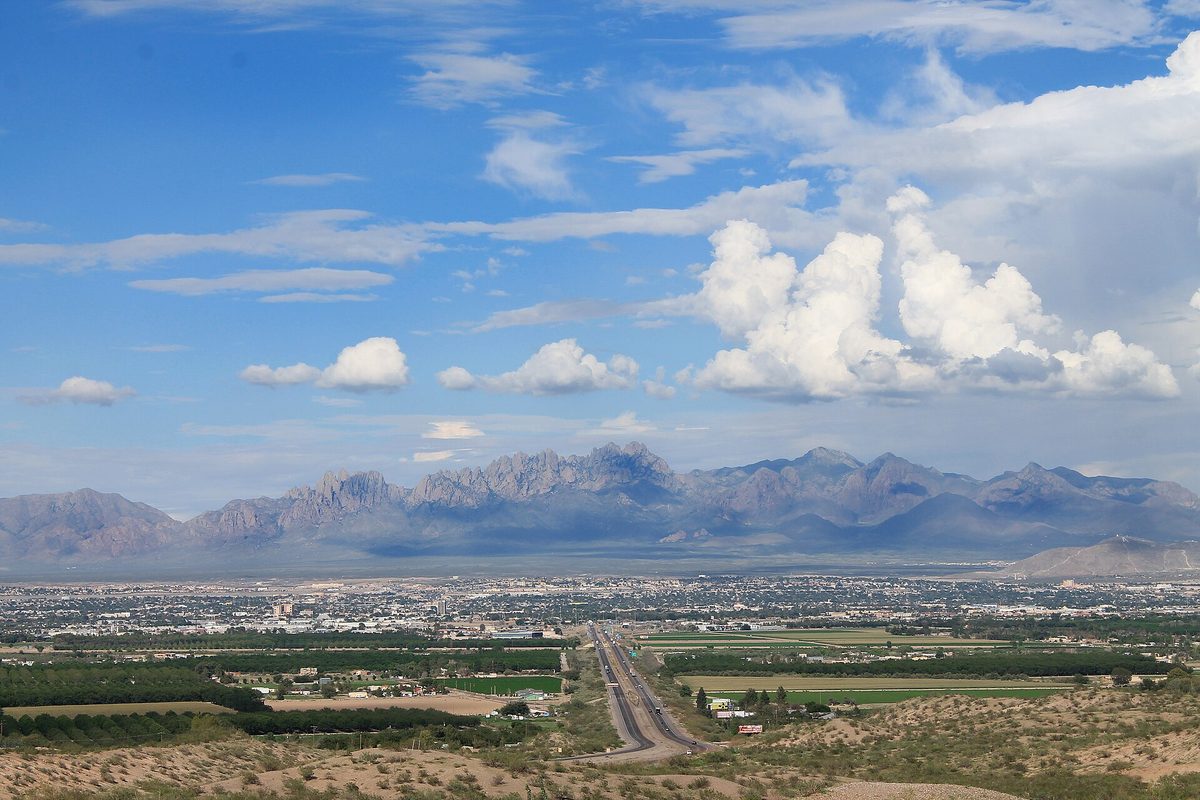 Las Cruces spread across the valley below the jagged Organ Mountains, with green fields and a highway in the foreground under big white clouds