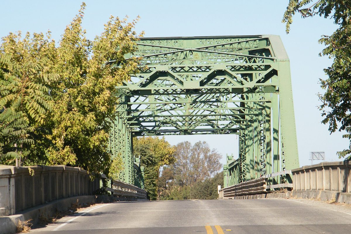 The green steel truss of the Manthey Road Bridge over the San Joaquin River in Lathrop, seen from the roadway with trees on both sides