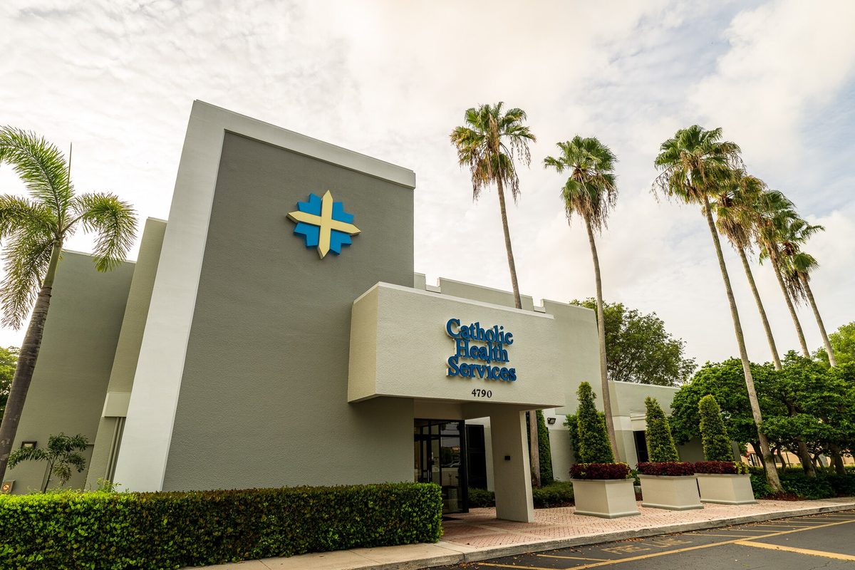 A gray Catholic Health Services office building with a blue and gold cross on its front, tall royal palms and trimmed hedges beside the parking lot on State Road 7 in Lauderdale Lakes