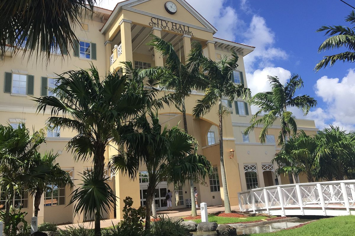 Lauderhill City Hall, a cream-colored building with a clock above the entrance, framed by palm trees and a white footbridge over a small pond