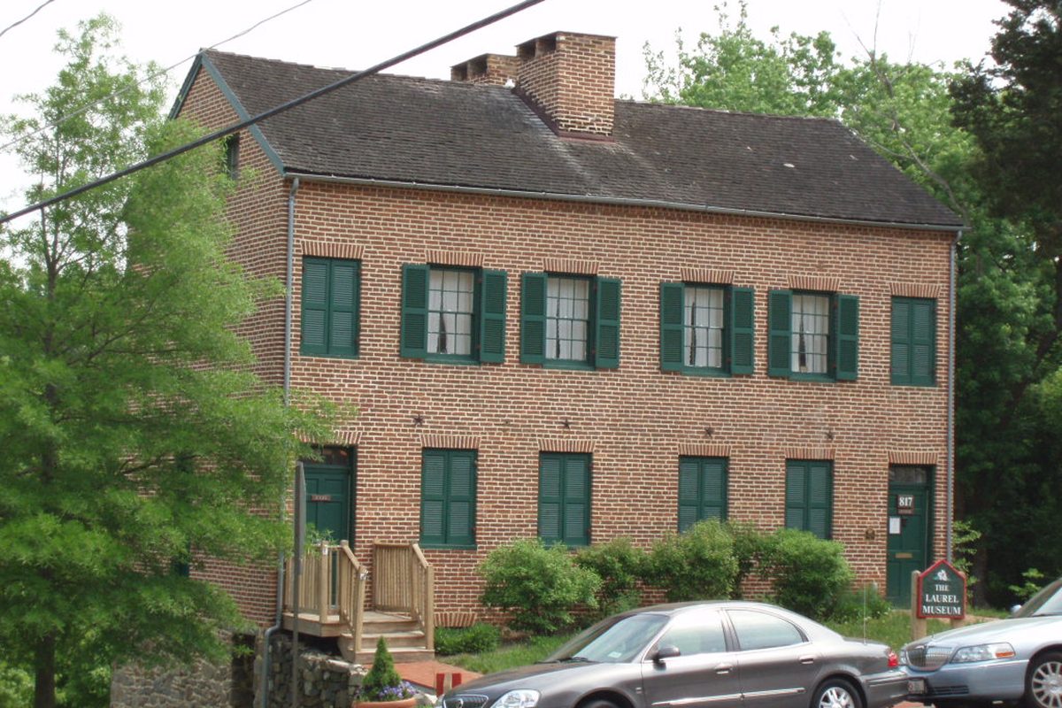 The Laurel Museum, a two-story red brick house with green shutters and chimneys, with two sedans parked out front