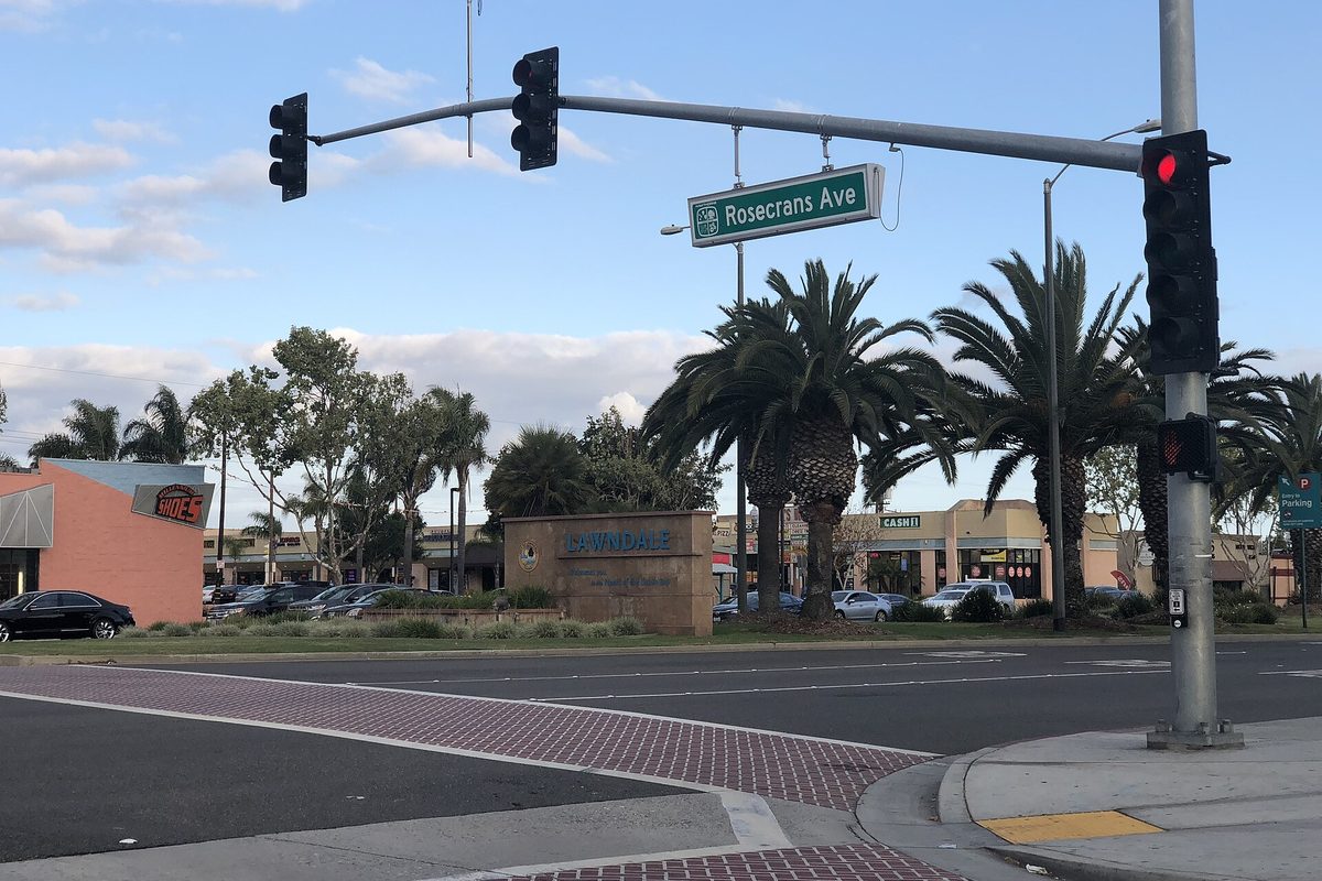 A Rosecrans Ave street sign and traffic signal over a Lawndale intersection, with palm trees and a stone Lawndale monument sign behind