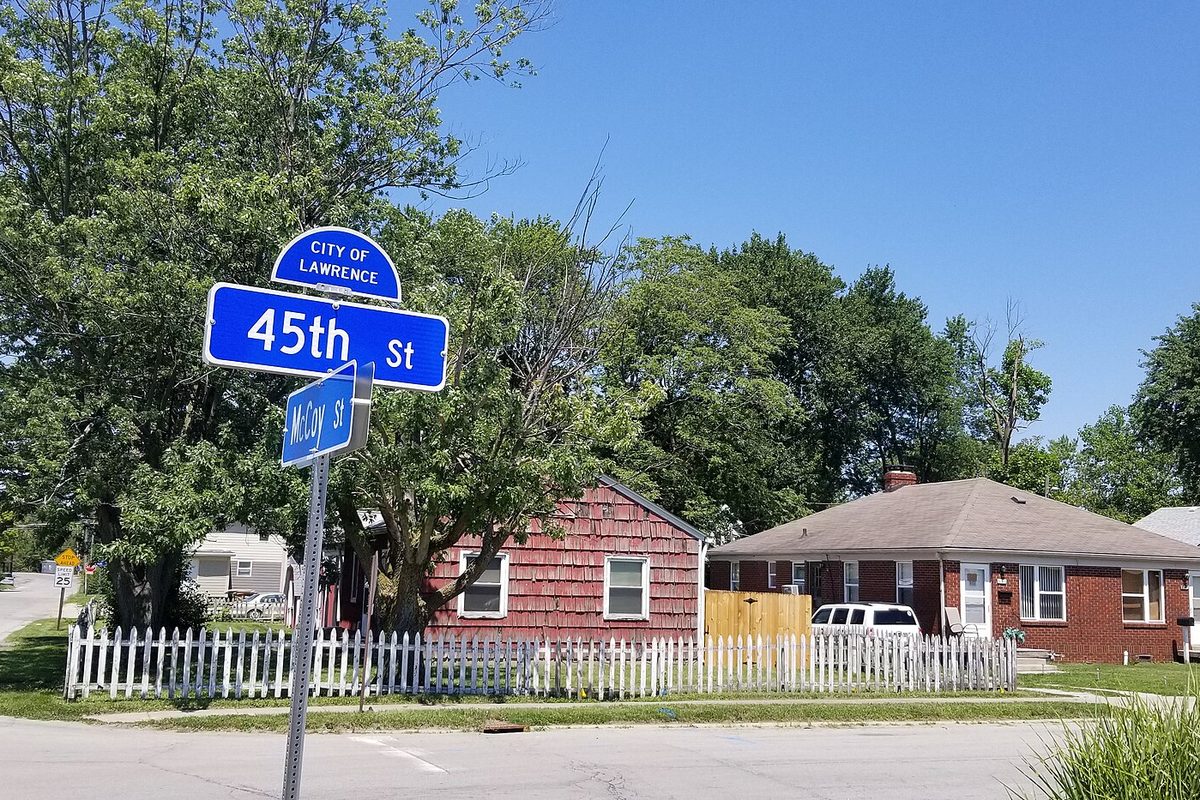 A blue City of Lawrence street sign at 45th Street and McCoy Street, with small houses behind a white picket fence and tall trees