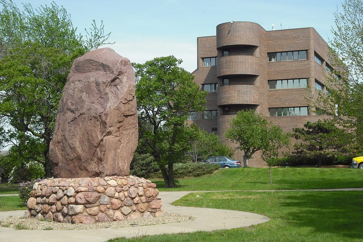 The large pink Shunganunga Boulder on a round stone base in front of Lawrence City Hall's brick building with its rounded corner tower