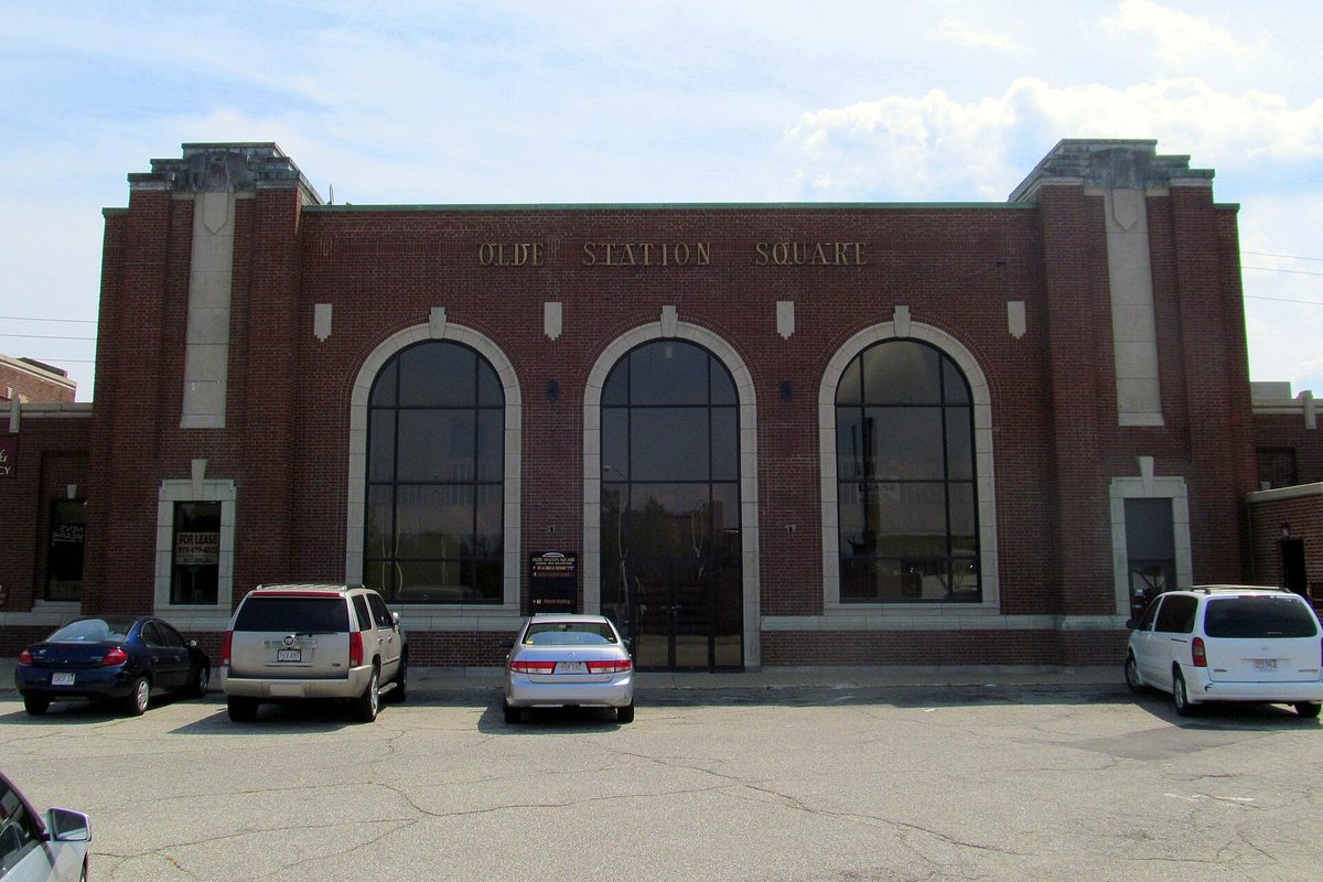 The brick Olde Station Square building in Lawrence, once the city's railroad station, with three tall arched windows and cars parked in front