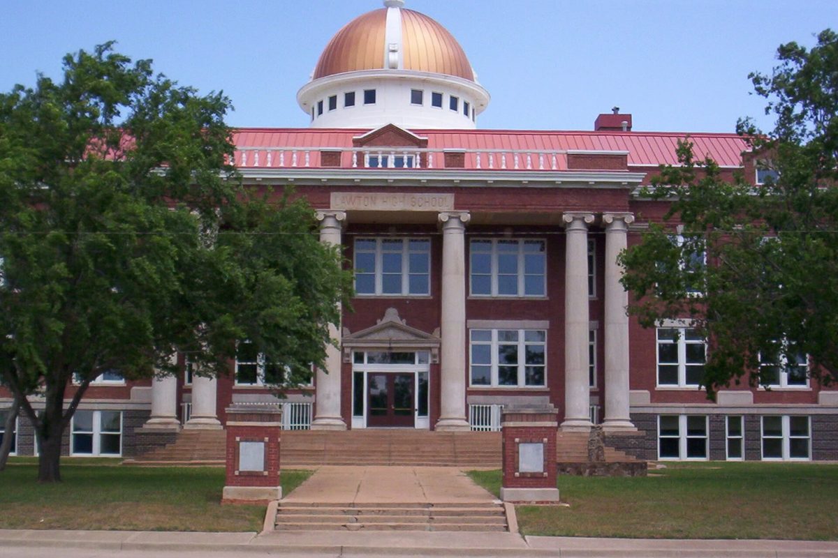 The old Lawton High School, a red-brick building with white columns at the entrance, a copper dome on top and trees on either side of the front walk