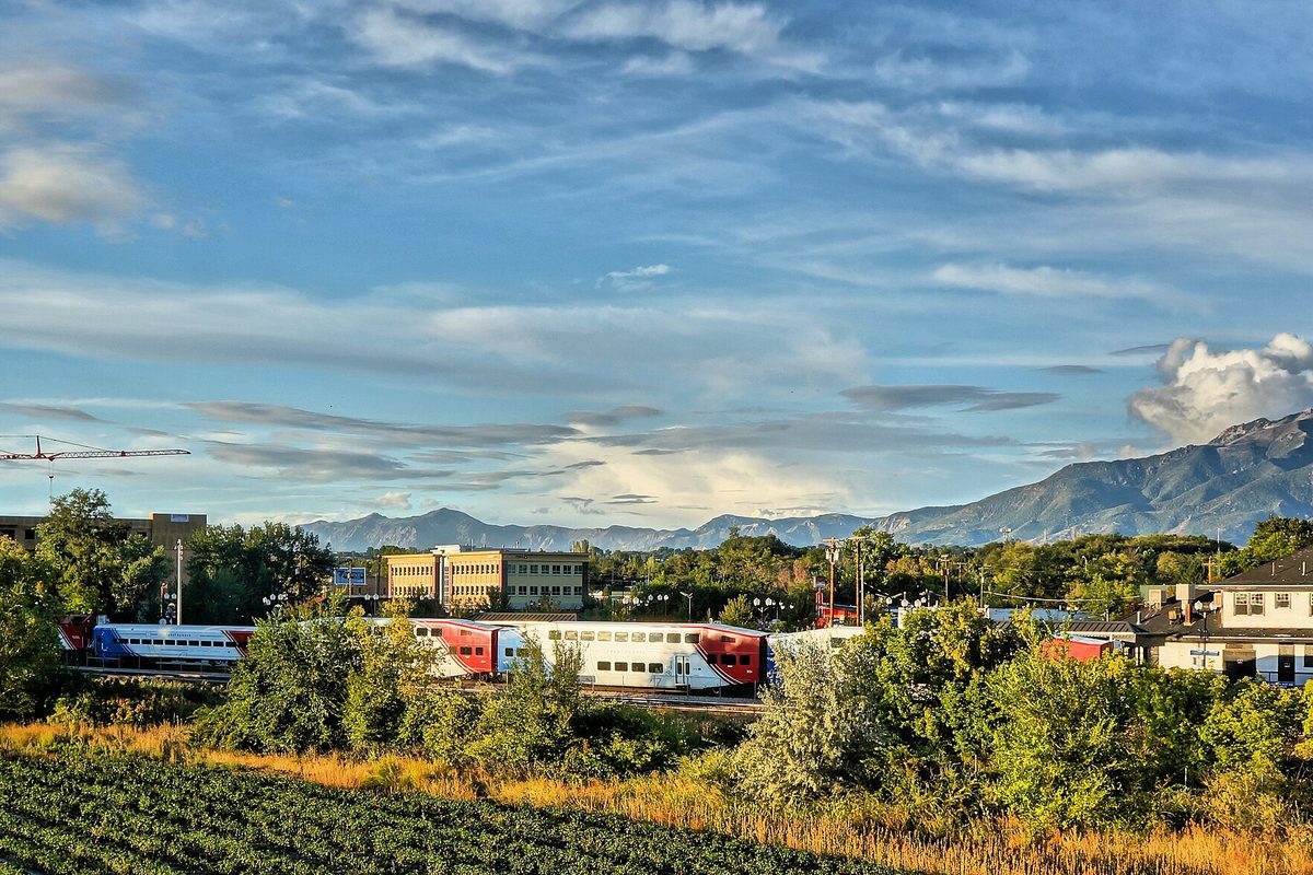 A FrontRunner commuter train passing through Layton beside a crop field, with low buildings, trees and mountains under a cloud-streaked sky