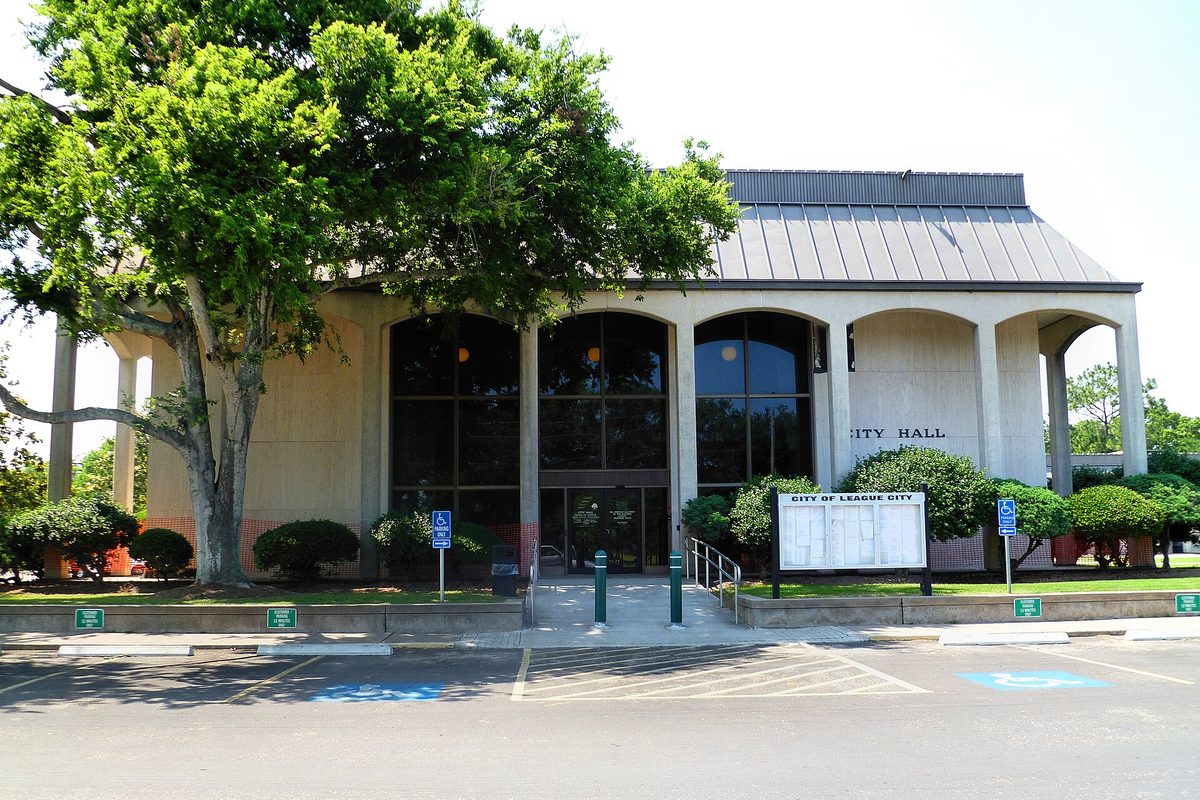 League City City Hall, a pale arched building with a metal roof and tall dark windows, shaded by a leafy tree beside the front walk and parking lot