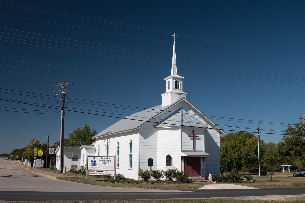Leander Presbyterian Church, a white wooden church with a tall steeple and a red cross over the door, beside a quiet road under power lines and a deep blue sky