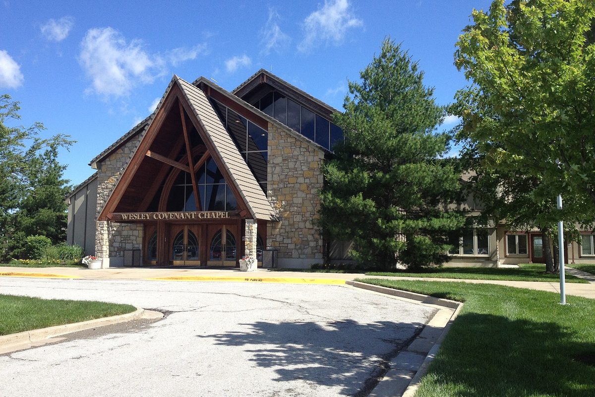 The stone and timber entrance of Wesley Covenant Chapel with its steep pitched roof, framed by tall evergreens under a blue sky