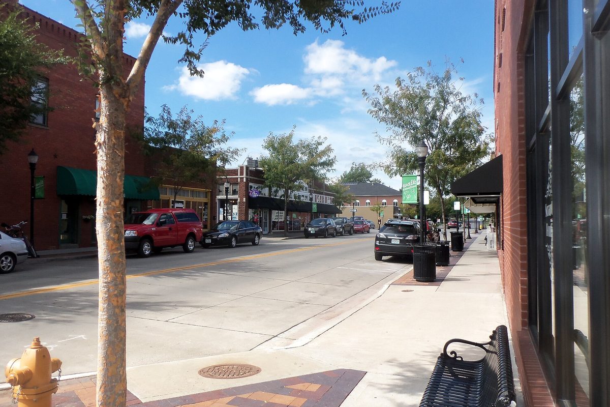 Downtown Lee's Summit on a sunny day: brick storefronts with green awnings, cars parked along the curb, a bench and a yellow fire hydrant