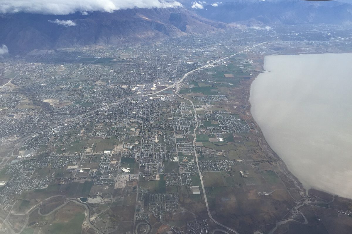 Aerial view of Lehi and the cities beside it along Interstate 15, with the shore of Utah Lake on the right and mountains under cloud behind