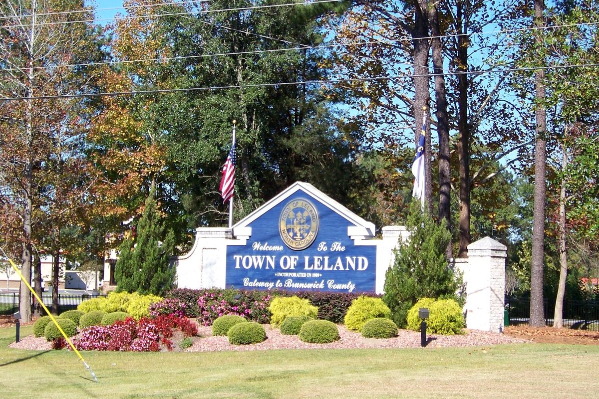 A blue Welcome to the Town of Leland sign reading Gateway to Brunswick County, with flags, trimmed shrubs and red flowers in front of tall pines