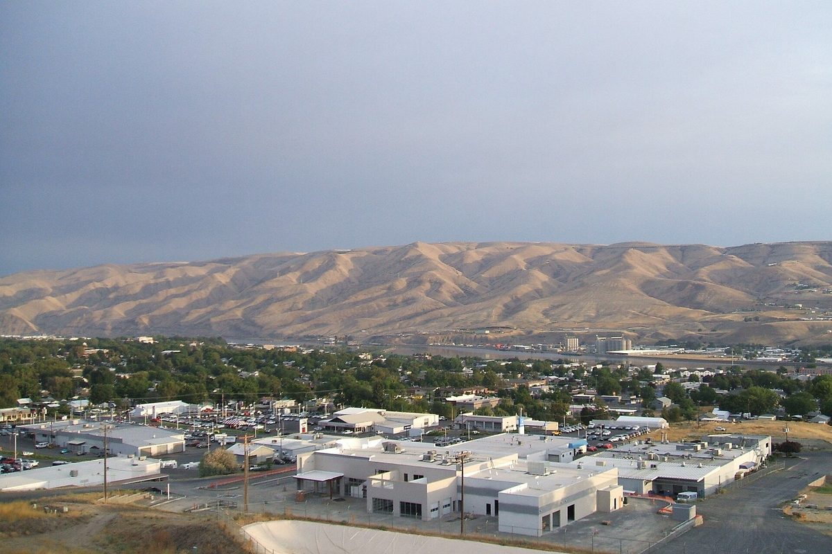Lewiston rooftops, shops and parking lots in the foreground, with bare golden hills rising beyond the Clearwater River under a gray sky