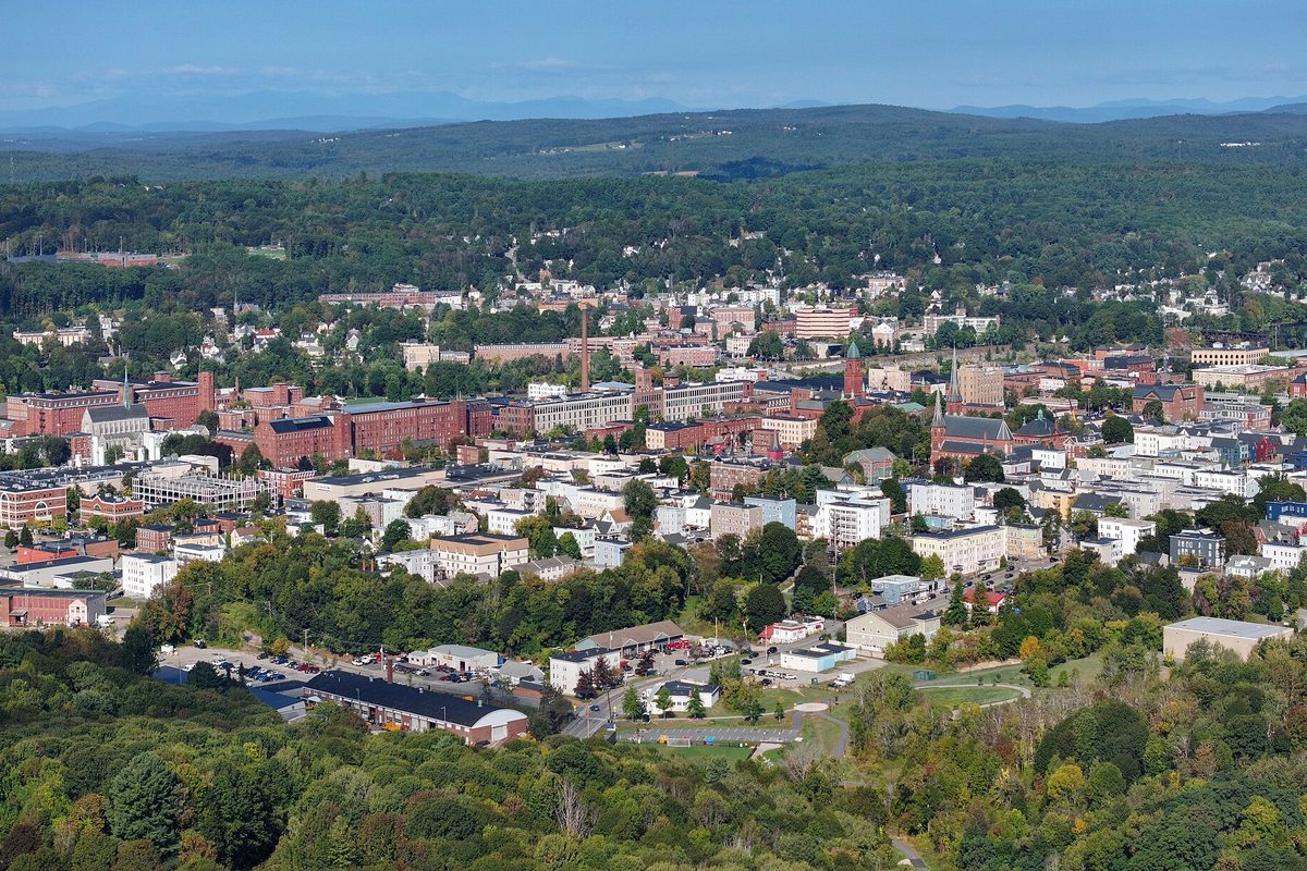 Aerial view of Lewiston in summer, red brick mill buildings and church spires among green trees, with low mountains on the horizon