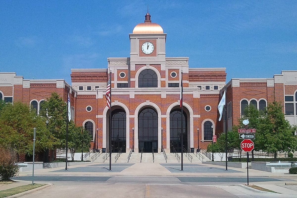 Lewisville City Hall, a red brick building with three tall arched doorways and a copper-domed clock tower, behind a stop sign at Church Street