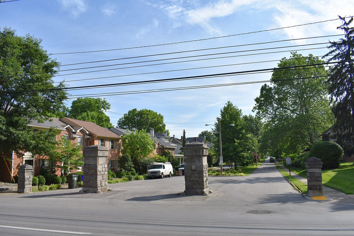 Stone gate pillars at the entrance to Mentelle Park in Lexington, a tree-lined street of brick houses beneath power lines on a sunny day