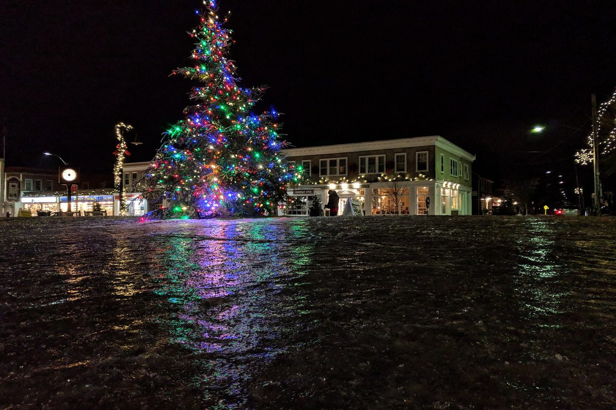 A tall Christmas tree strung with colored lights in Depot Square, Lexington, at night, its glow reflected on wet pavement in front of storefronts