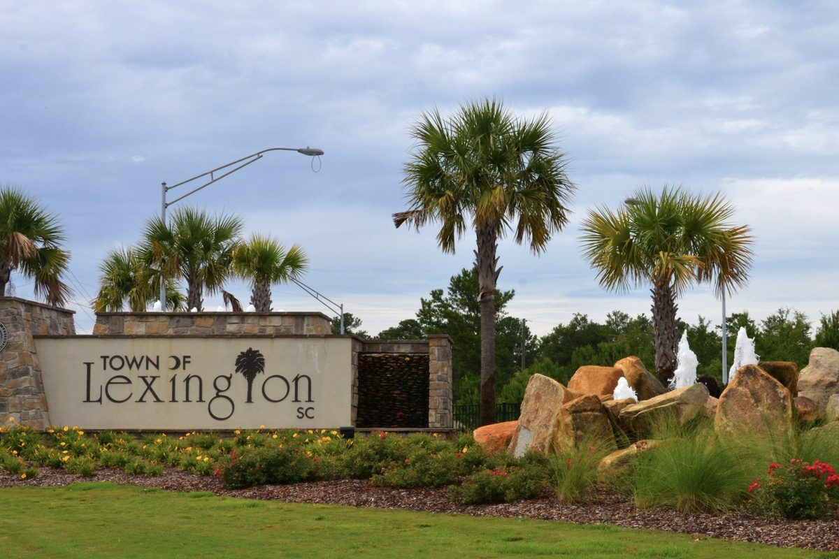 A stone Town of Lexington SC welcome sign with a palmetto emblem, flanked by palmetto trees, flowers, large boulders and small fountains
