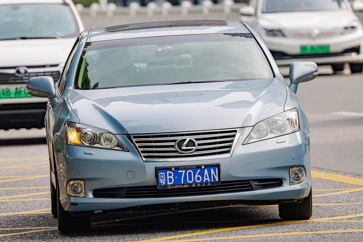 A light blue Lexus ES sedan seen head-on, driving in a lane with yellow road markings and white cars behind it