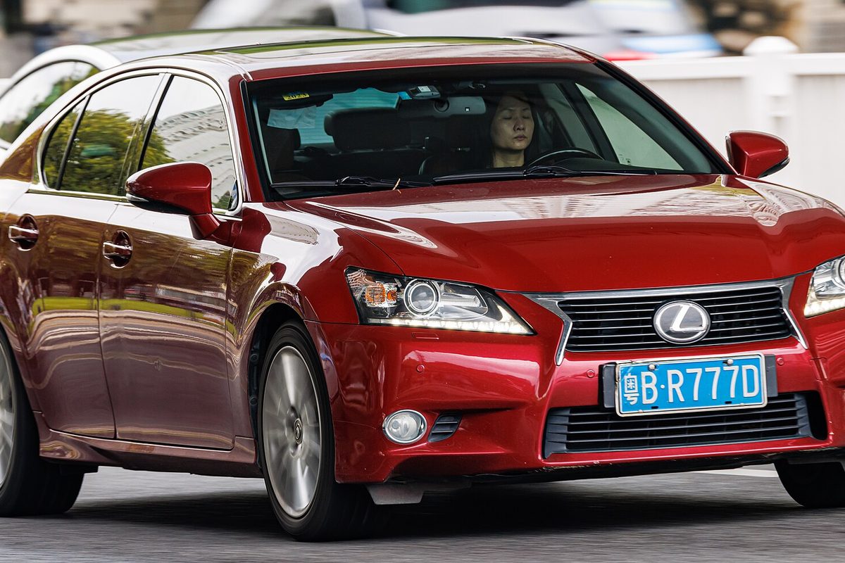 A red Lexus GS sedan driving past, seen from the front three-quarter, with a driver behind the wheel