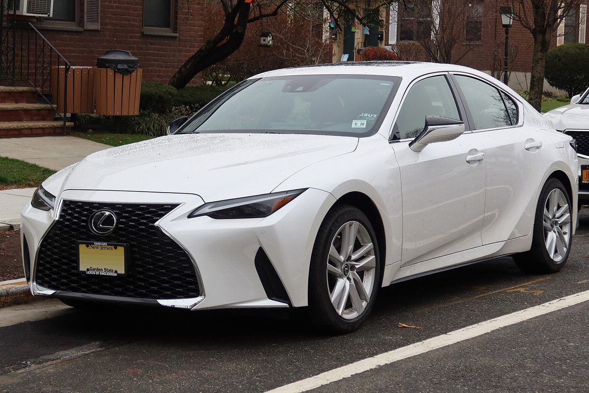 A white Lexus IS sedan with a black mesh grille, parked at a curb on a residential street beside brick row houses