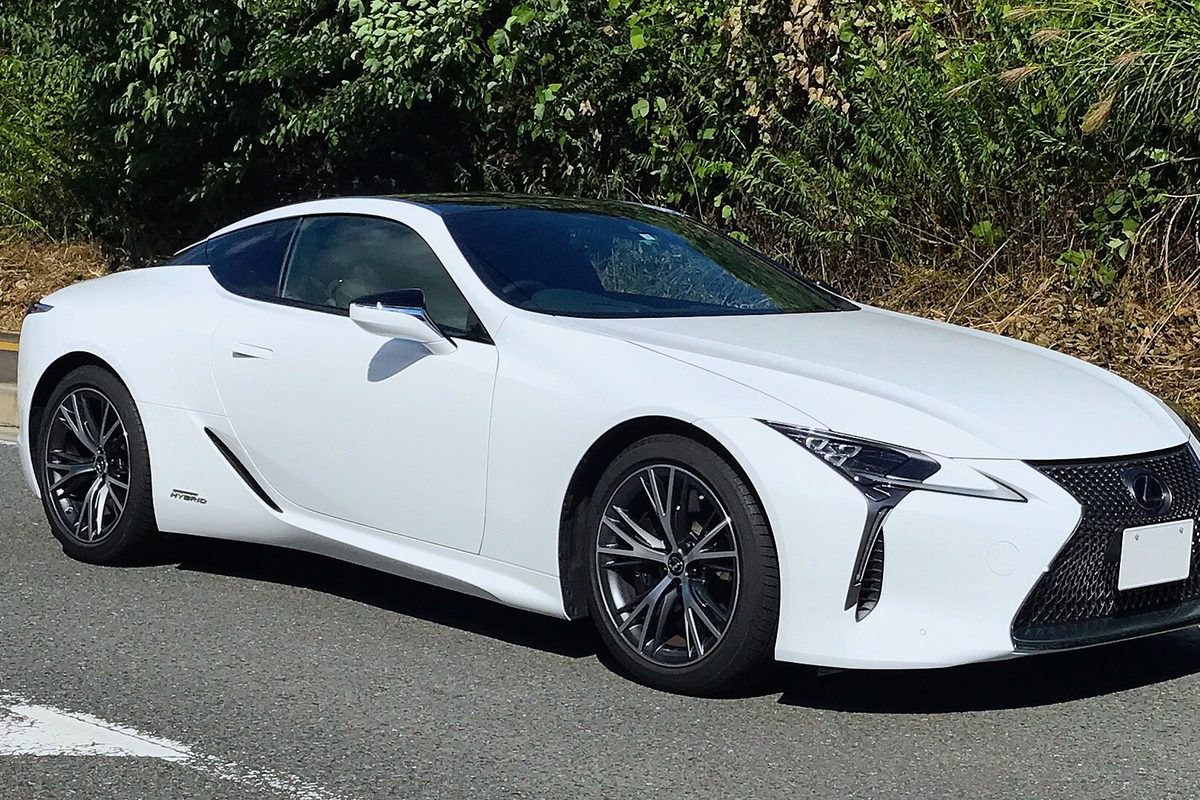 A white Lexus LC 500h coupe with dark wheels on a road, green bushes behind it