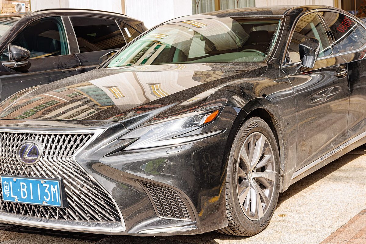 A black Lexus LS sedan with a large chrome mesh grille, parked beside a dark SUV, its paint reflecting the buildings around it
