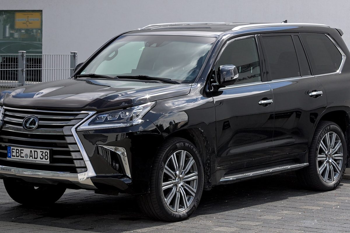 A black third-generation Lexus LX 570 with a chrome spindle grille and multi-spoke wheels, parked on paving stones beside a gray building