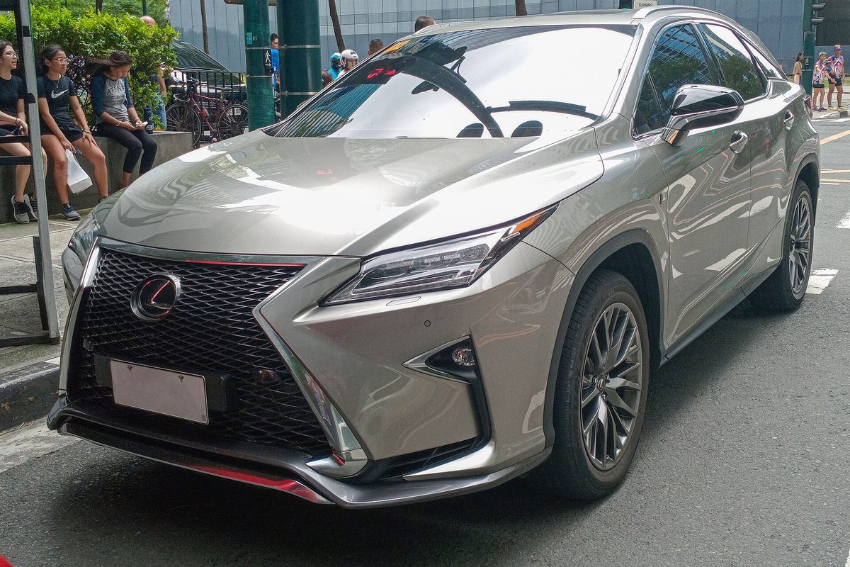 A champagne-silver Lexus RX F Sport SUV with a black mesh grille, parked on a city street with people on the sidewalk behind it