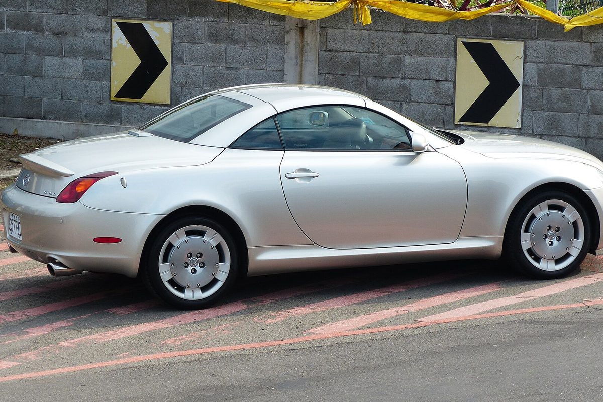 A silver second-generation Lexus SC 430 hardtop convertible with its roof up, parked beside a block wall with black-and-yellow chevron signs