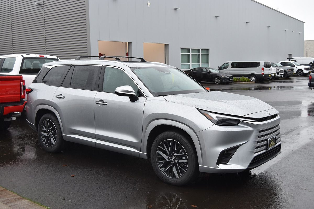 A silver 2024 Lexus TX 350 three-row SUV parked on a wet dealership lot next to a red pickup