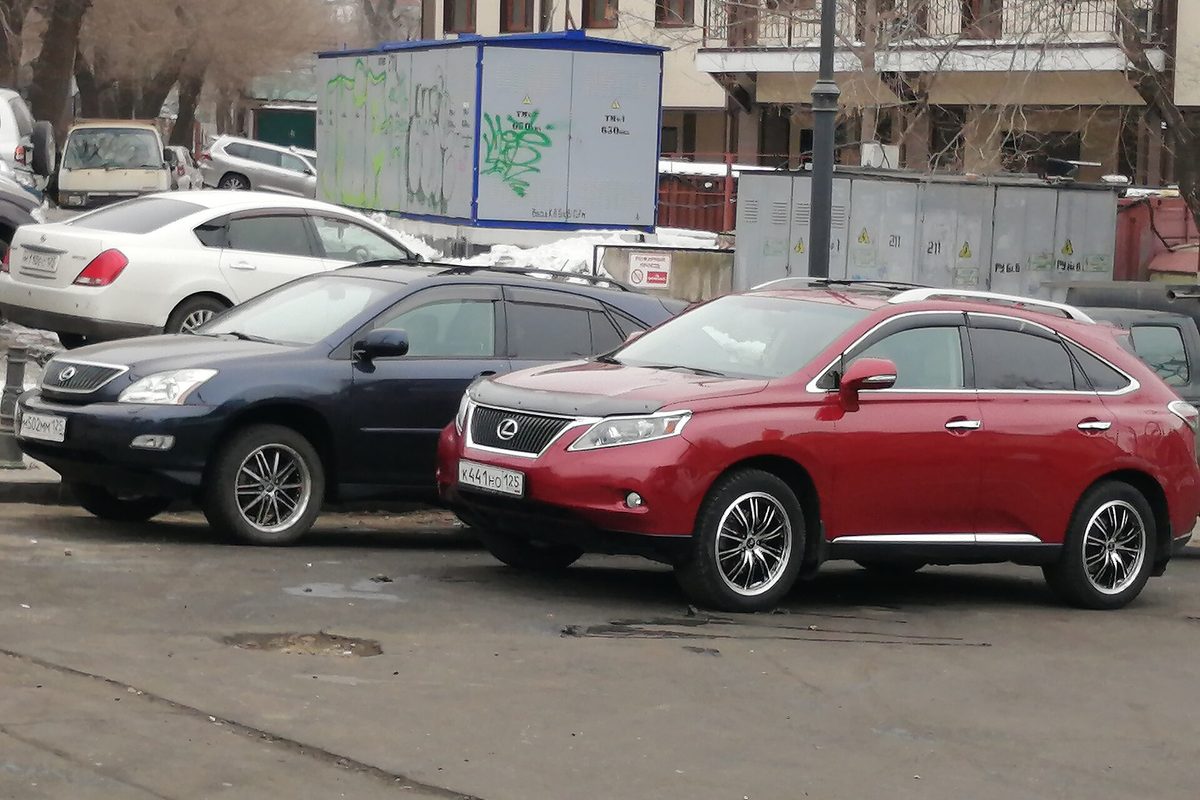 A red 2009-2012 Lexus RX parked next to a dark blue 2006-2009 Lexus RX on a city street in Vladivostok, Russia, with snow piled behind them