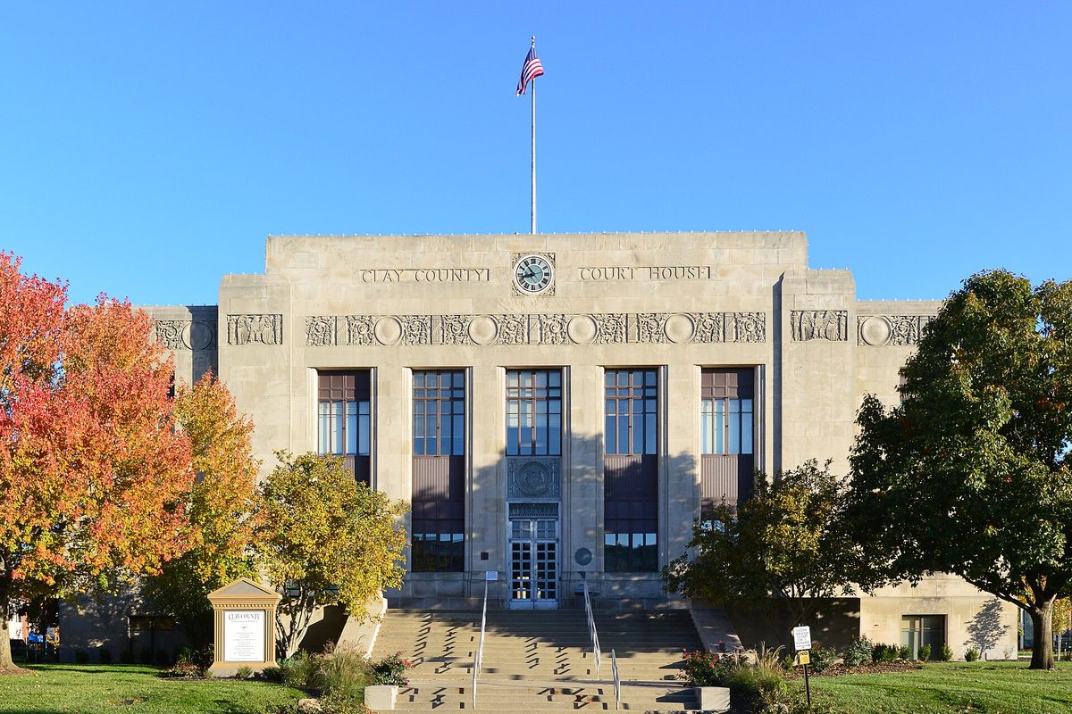 The pale stone Clay County Courthouse in Liberty, with a clock above the entrance, a flag on the roof and autumn trees on both sides