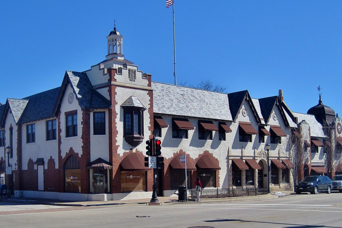 Libertyville's white stucco Public Service Building with brick trim, red awnings and a small cupola, on a downtown street corner under a blue sky