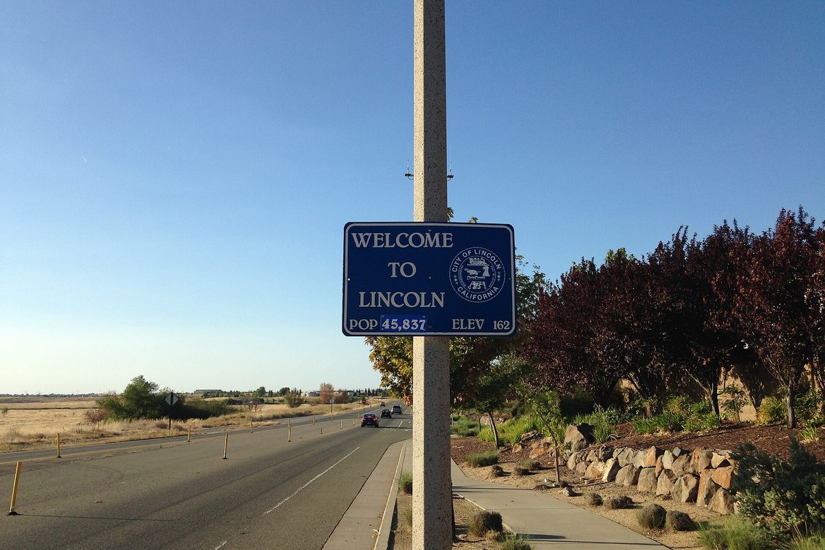 A blue Welcome to Lincoln sign on a concrete pole beside a road, with a sidewalk, red-leafed trees and open fields under a clear sky