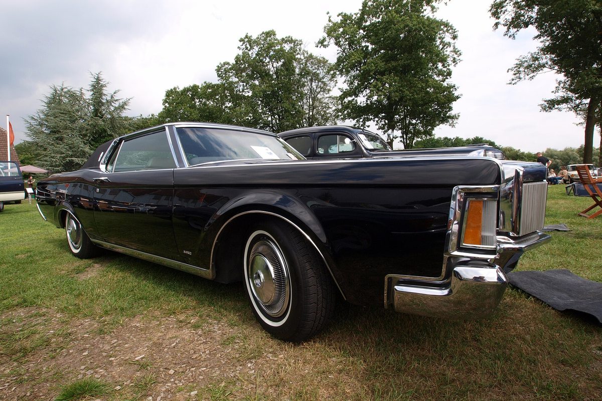 A black two-door Lincoln Continental with whitewall tires and chrome bumpers, parked on grass at an outdoor classic car show